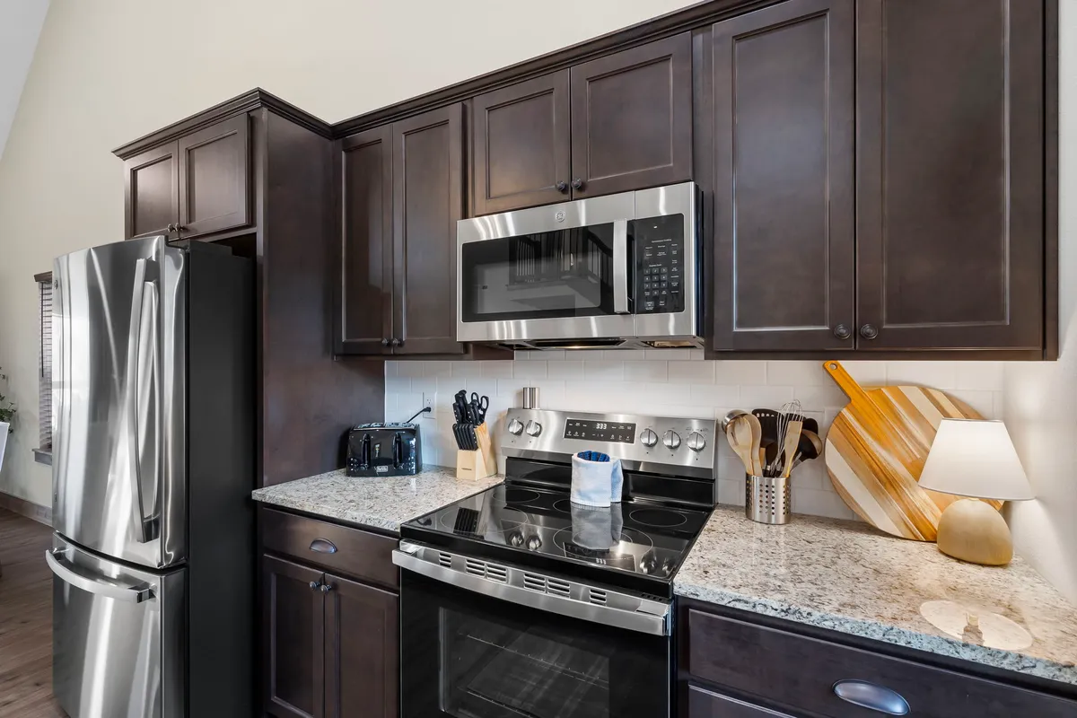 A contemporary kitchen with stainless steel appliances, granite countertops, and cozy modern cabinetry. A decorative cutting board and table lamp add warmth and personality to the space, emphasizing a mix of function and comfort.