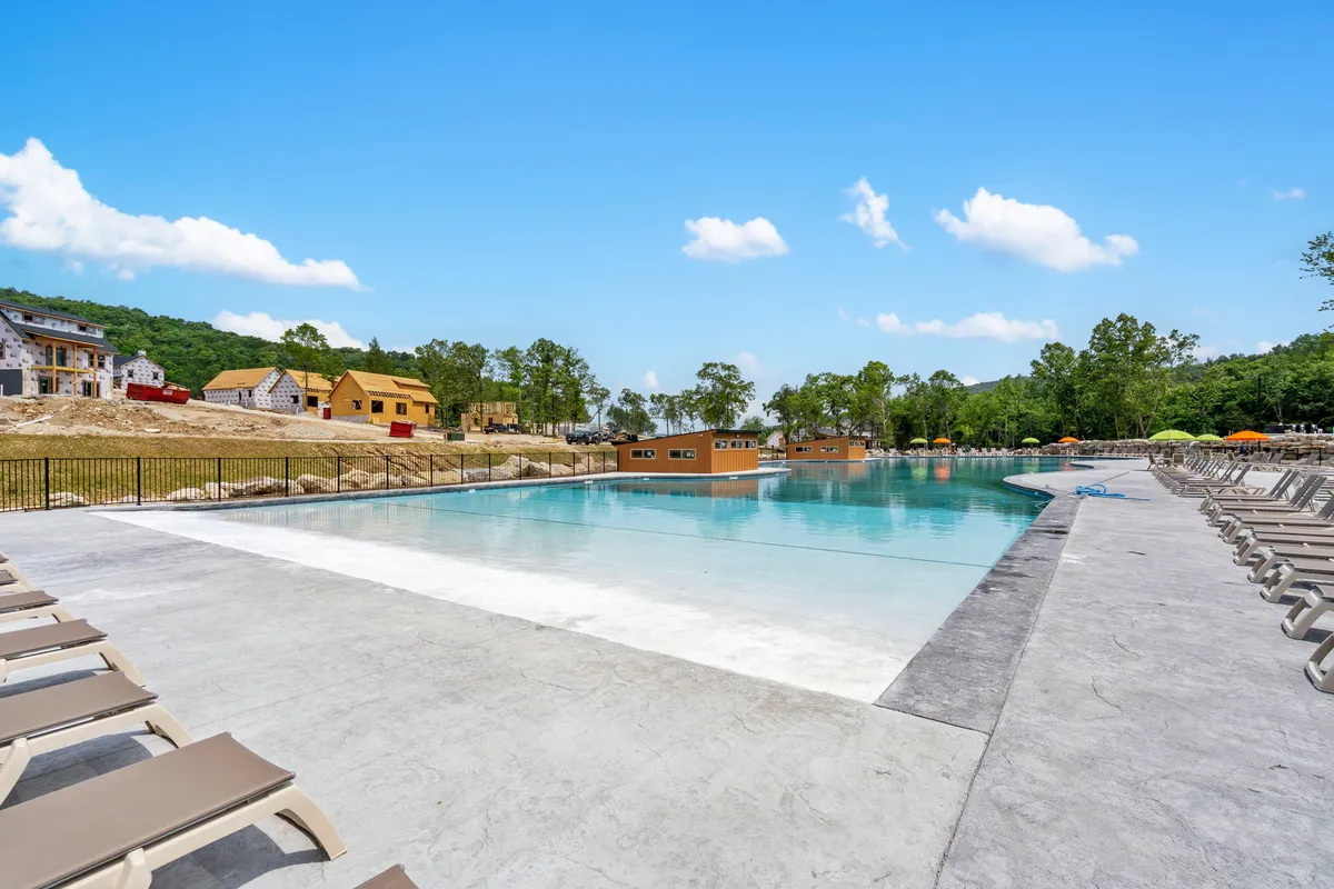 An outdoor swimming pool is the focal point, surrounded by colorful umbrellas and lounge chairs. The background shows resort buildings, and the overall mood is leisure-oriented and cheerful, with space for families to swim, socialize, and relax by the water.