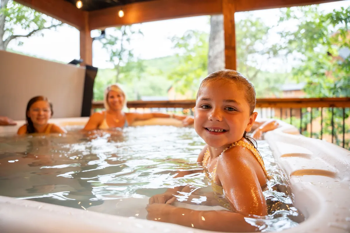 A smiling child in the foreground enjoys relaxing in an outdoor hot tub, while two women share a joyful moment in the background. The hot tub is nestled within a natural setting, surrounded by trees and open air, creating a sense of relaxation after outdoor adventures.