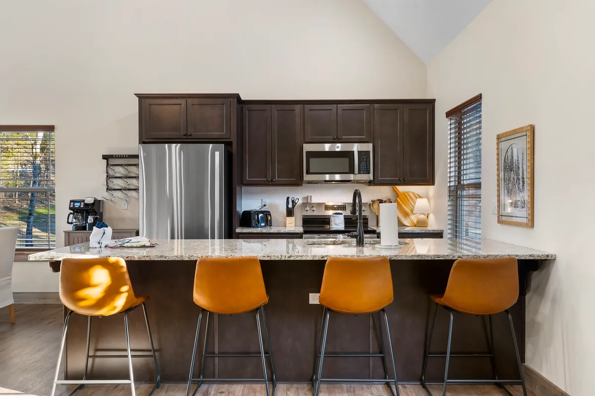 A bright, modern kitchen with granite countertops, stainless steel appliances, bar stools at a breakfast bar, and large windows letting in natural light. The setting feels inviting and functional for both cooking and casual dining.