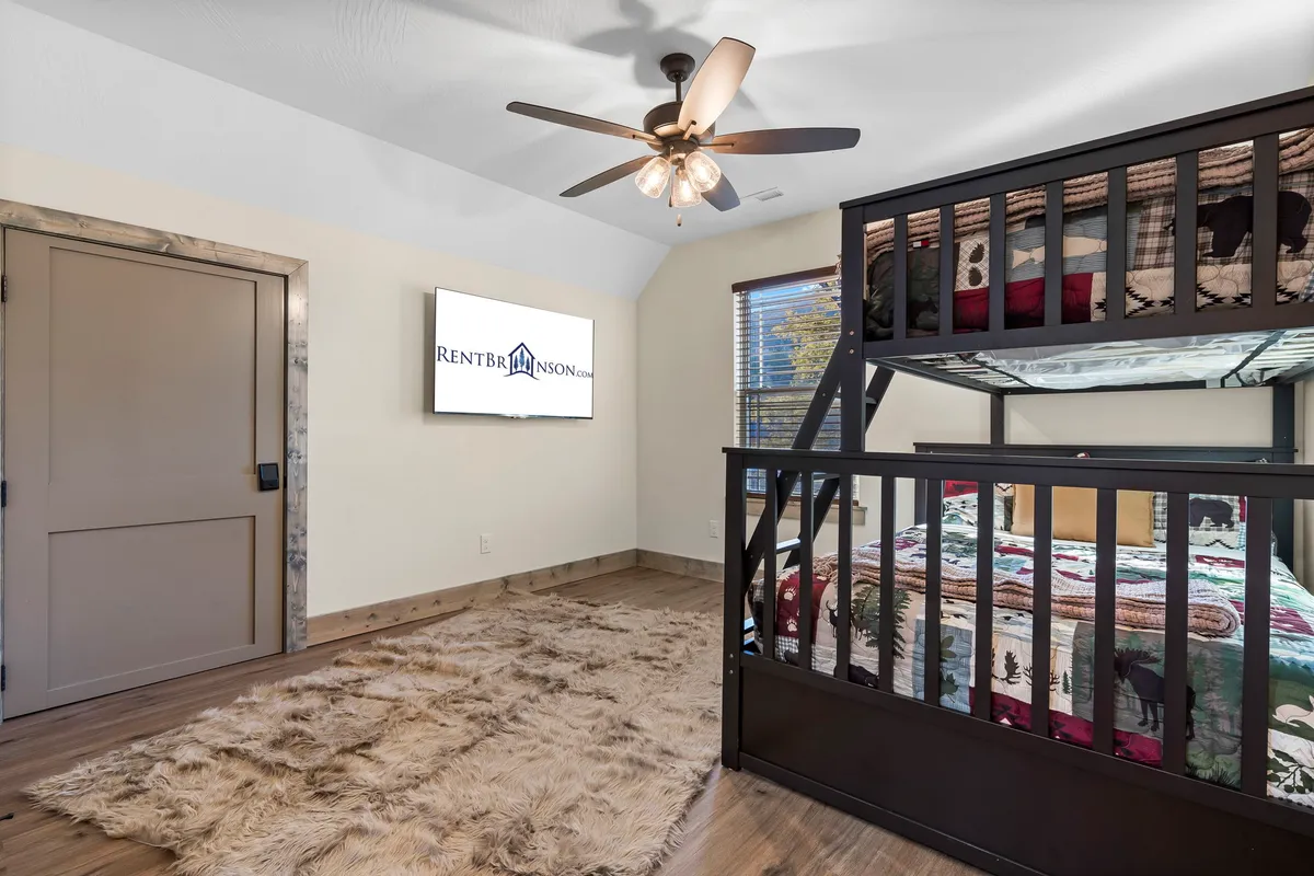 A playful bunk room featuring a twin over full bunk bed adorned with a decorative quilt, a cozy rug, ceiling fan, and a TV. The design is family-friendly and whimsical, ideal for kids or teens seeking their own fun space.