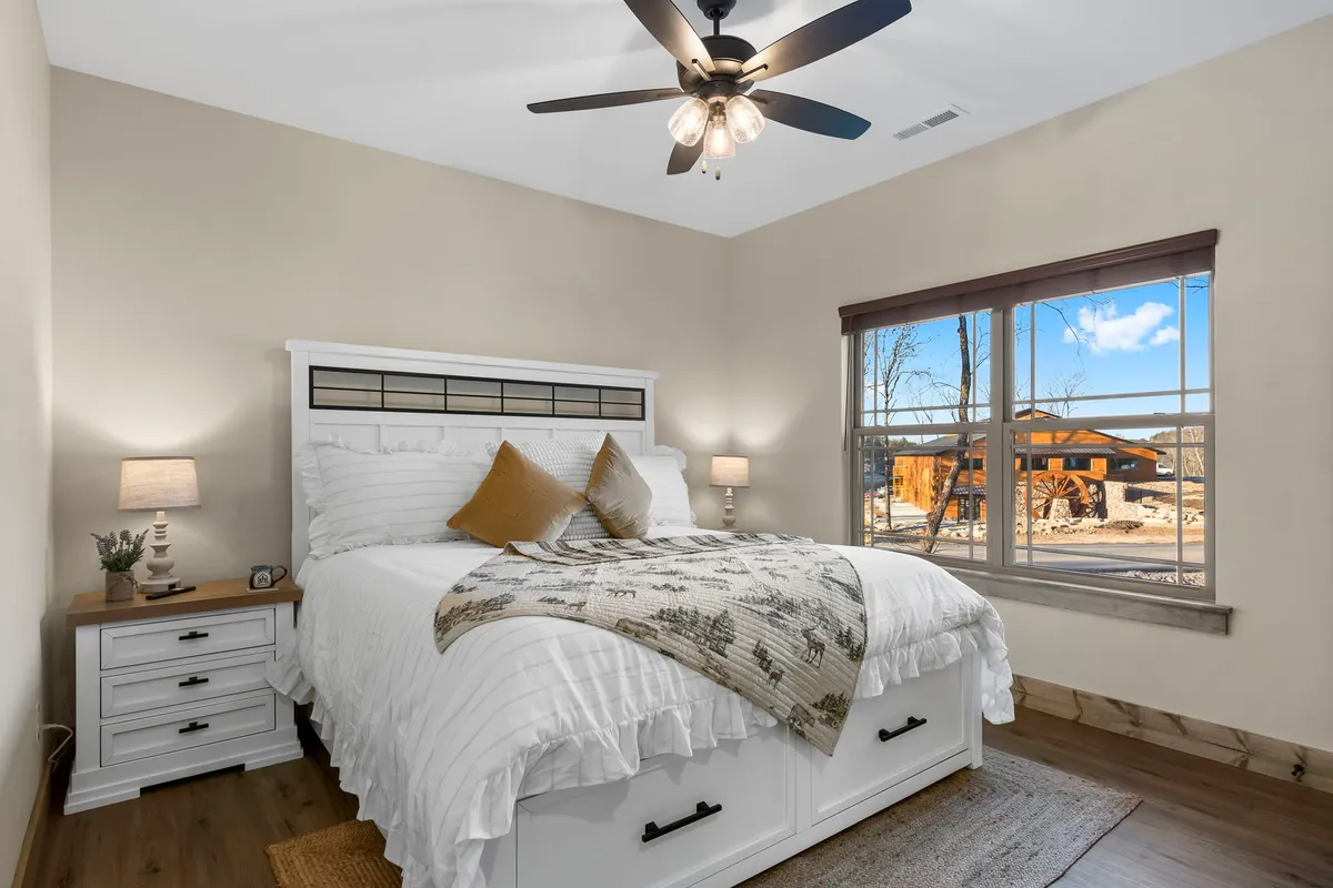 A stylish bedroom furnished with a neatly made bed, a window letting in morning light, a nightstand, a lamp, and a ceiling fan. The room exudes warmth and comfort, ideal for unwinding or starting the day refreshed.