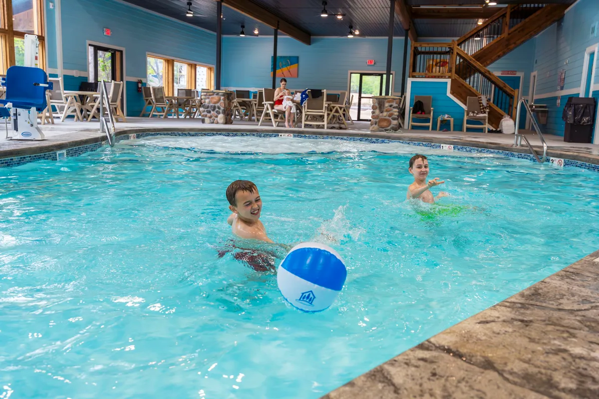 This interior shot spotlights the year-round indoor swimming pool, with children playing, a beach ball, lounge chairs, and abundant natural light. The mood is cheerful and energetic, emphasizing family enjoyment and leisure.