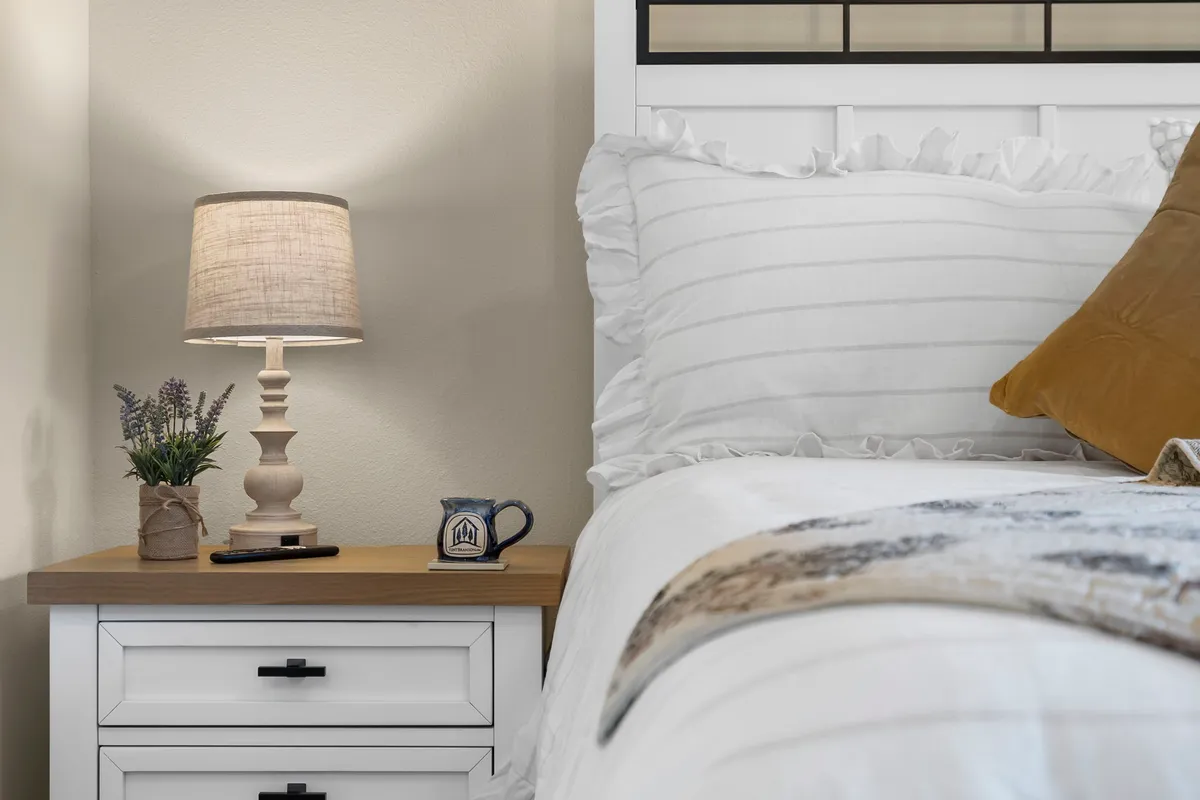 A serene bedroom featuring a bed with crisp white bedding, a nightstand with a warm lamp, a decorative pillow, and a small plant. Soft natural light filters in, creating a peaceful and inviting atmosphere perfect for relaxation and restful sleep.