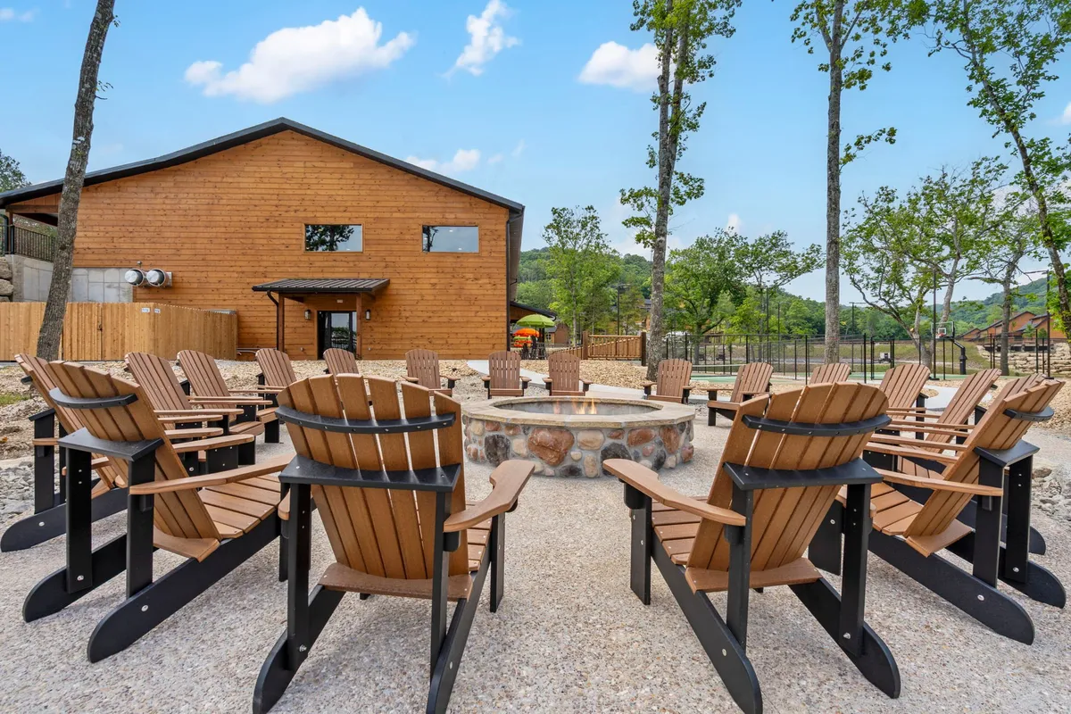 This image showcases a relaxed outdoor gathering space featuring a fire pit surrounded by Adirondack chairs and a wooden building set amidst leafy trees. The setting is ideal for storytelling, grilling, and unwinding in the evening, with the warm glow of sunset visible beyond the trees.