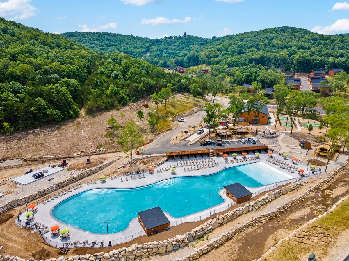 The photo features a large, sparkling outdoor swimming pool surrounded by sun loungers, lush greenery, and a wooden deck area. The scene is lively yet peaceful, suggesting family fun and relaxation under the sun.
