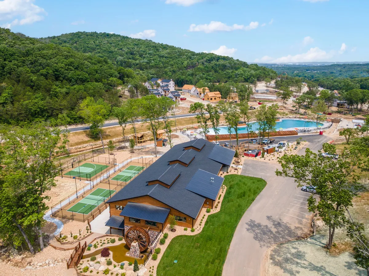 This image displays the vibrant community amenities area, with a community center building, a sparkling swimming pool, tennis courts, landscaped gardens, and the gentle rise of surrounding hills. The scene buzzes with the possibility of fun—swimming, sports, and community events—rain or shine.