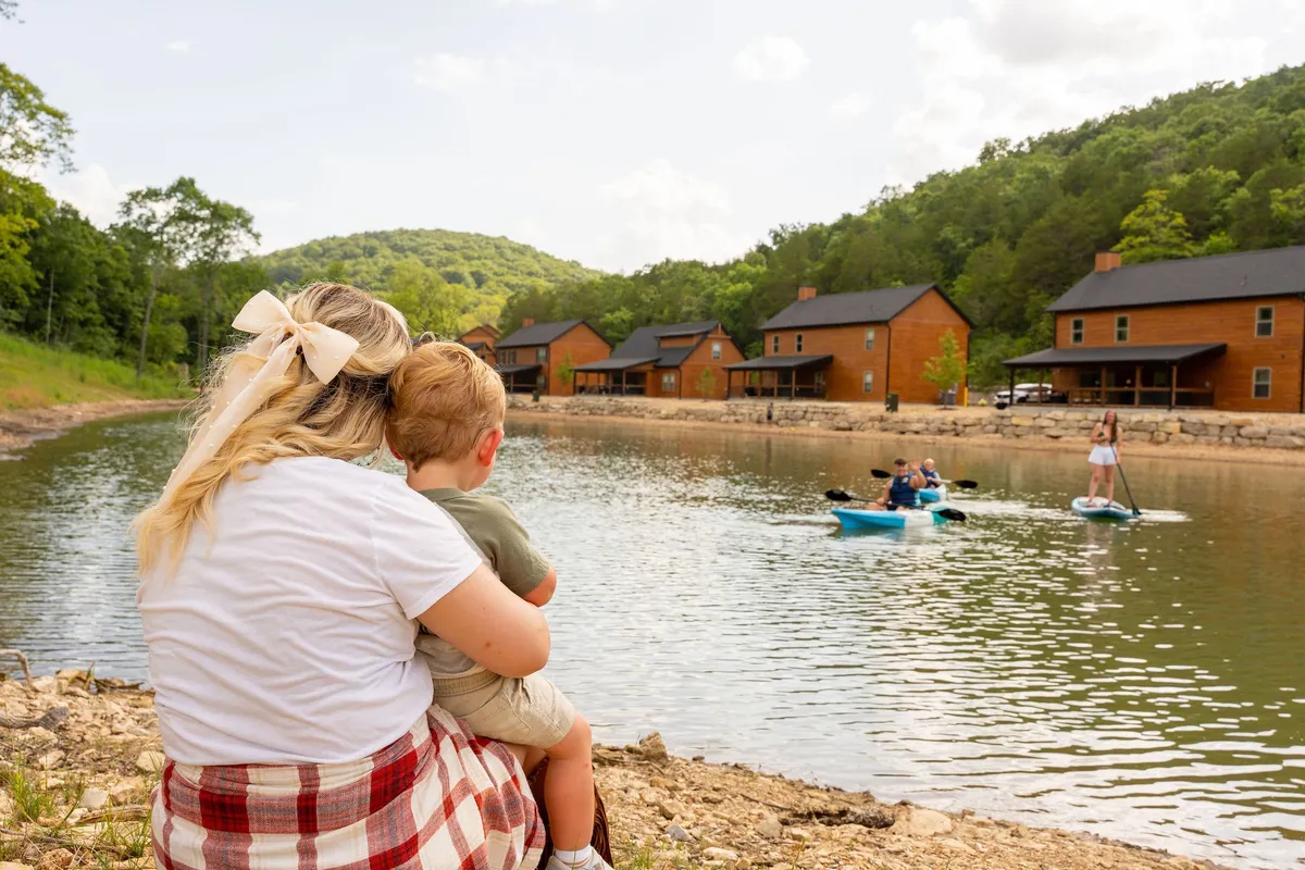 A mother and child enjoy quiet moments together on the lakefront, with wooden cabins nestled by the water’s edge and people kayaking in the background. The scene is surrounded by lush greenery and a peaceful, reflective surface of the lake.